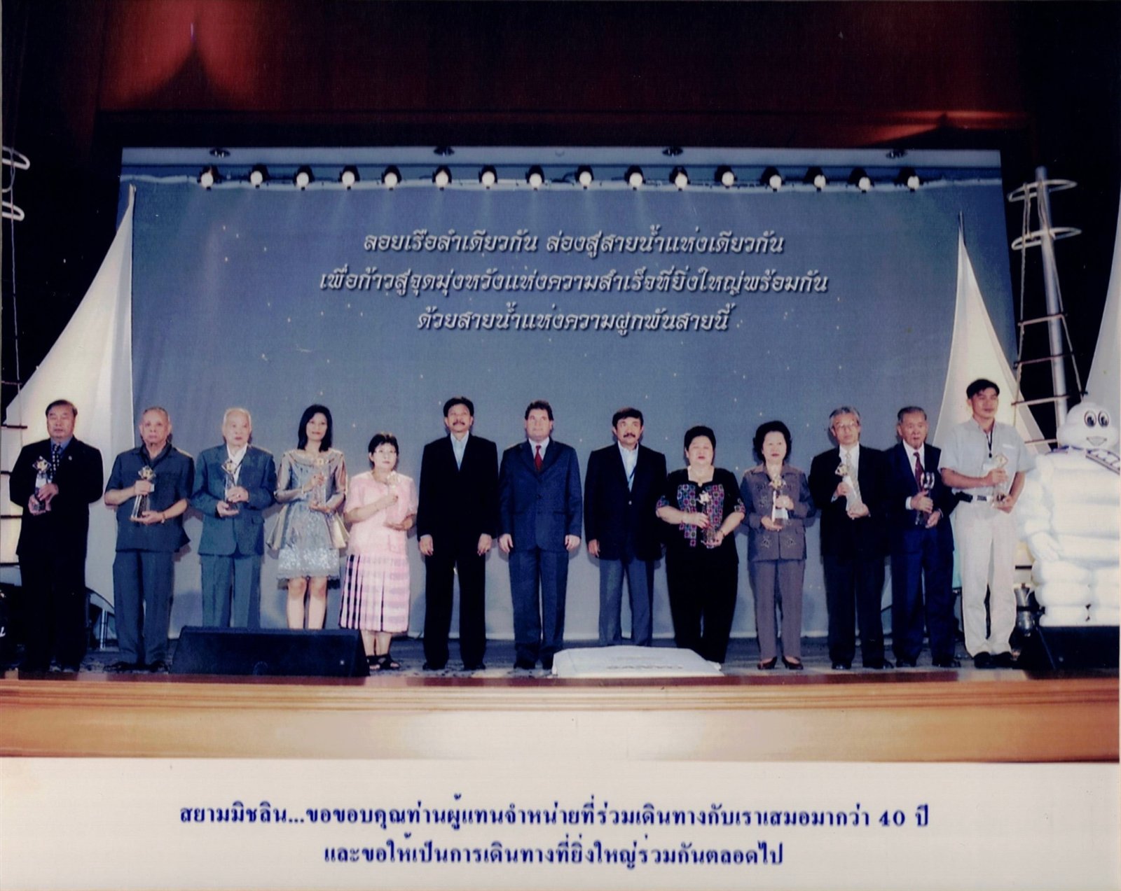งานขอบคุณผู้แทนจำหน่ายของสยามมิชลิน ผู้แทนจำหน่ายถือรางวัลบนเวที ใต้ข้อความ ลอยเรือลำเดียวกัน ล่องสู่สายน้ำแห่งเดียวกัน พร้อมคำขอบคุณที่ร่วมเดินทางกันมากว่า 40 ปี